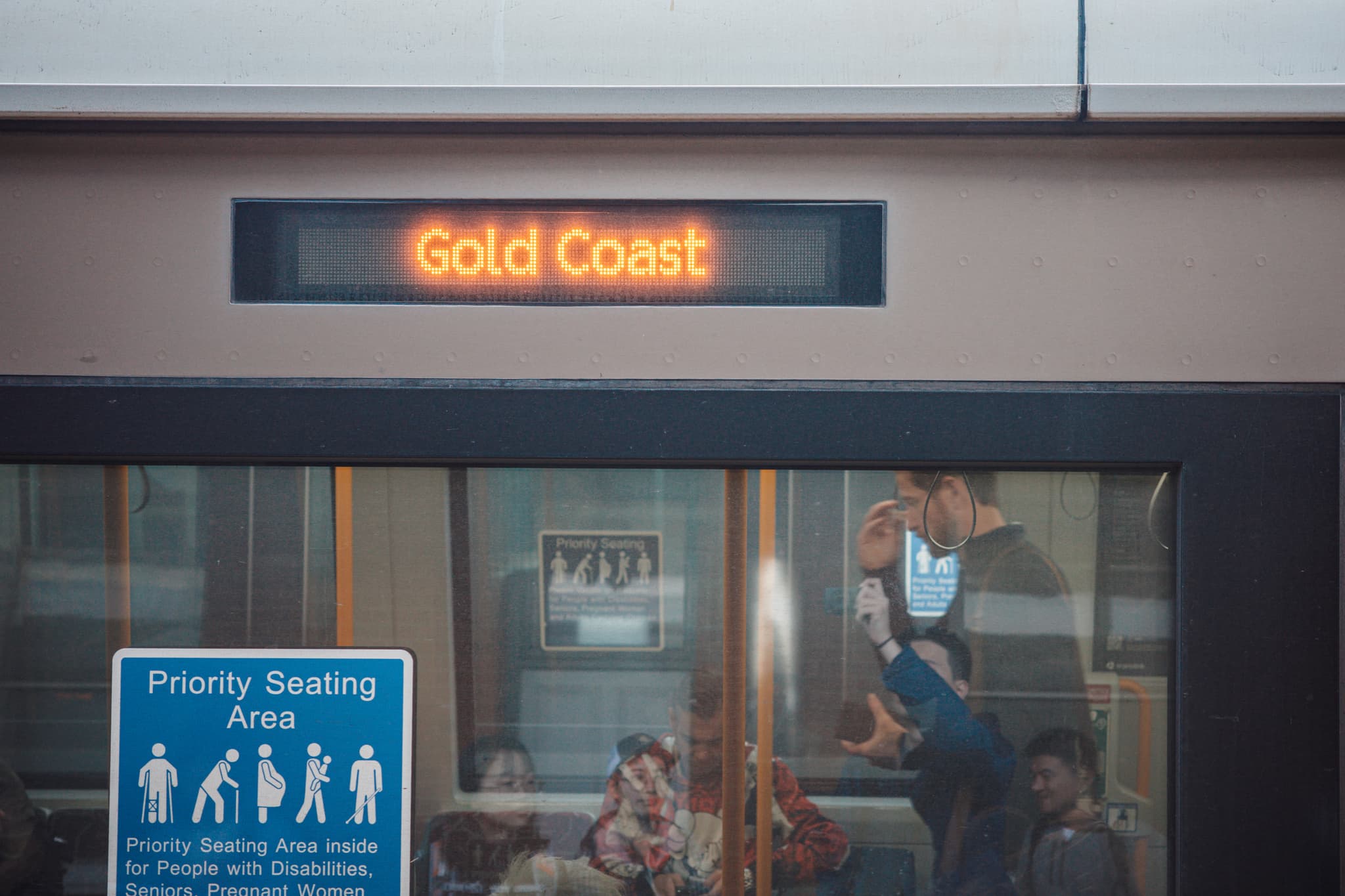 Train platform scene with an electronic departure board reading “Gold Coast” above a windowed waiting area and a priority seating sign on the left