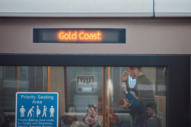 Train platform scene with an electronic departure board reading “Gold Coast” above a windowed waiting area and a priority seating sign on the left