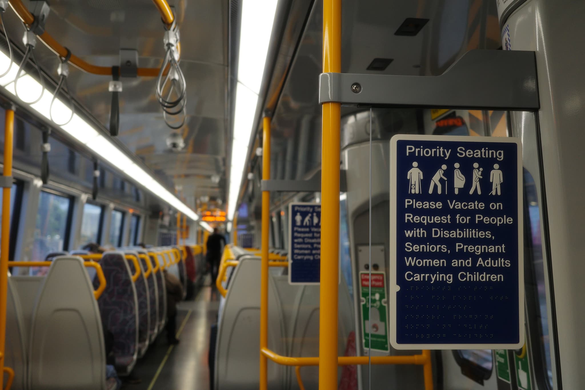 Interior of a passenger train with rows of seats, yellow handrails, and a priority seating sign near the door