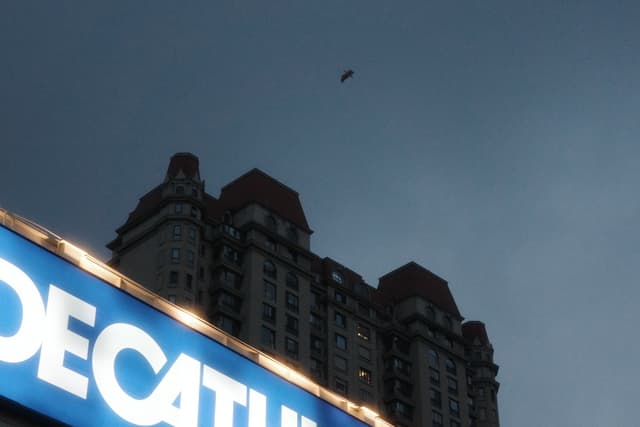A tall building with red rooftops under a dark, cloudy sky, with a glowing sign in the foreground and a small object flying above