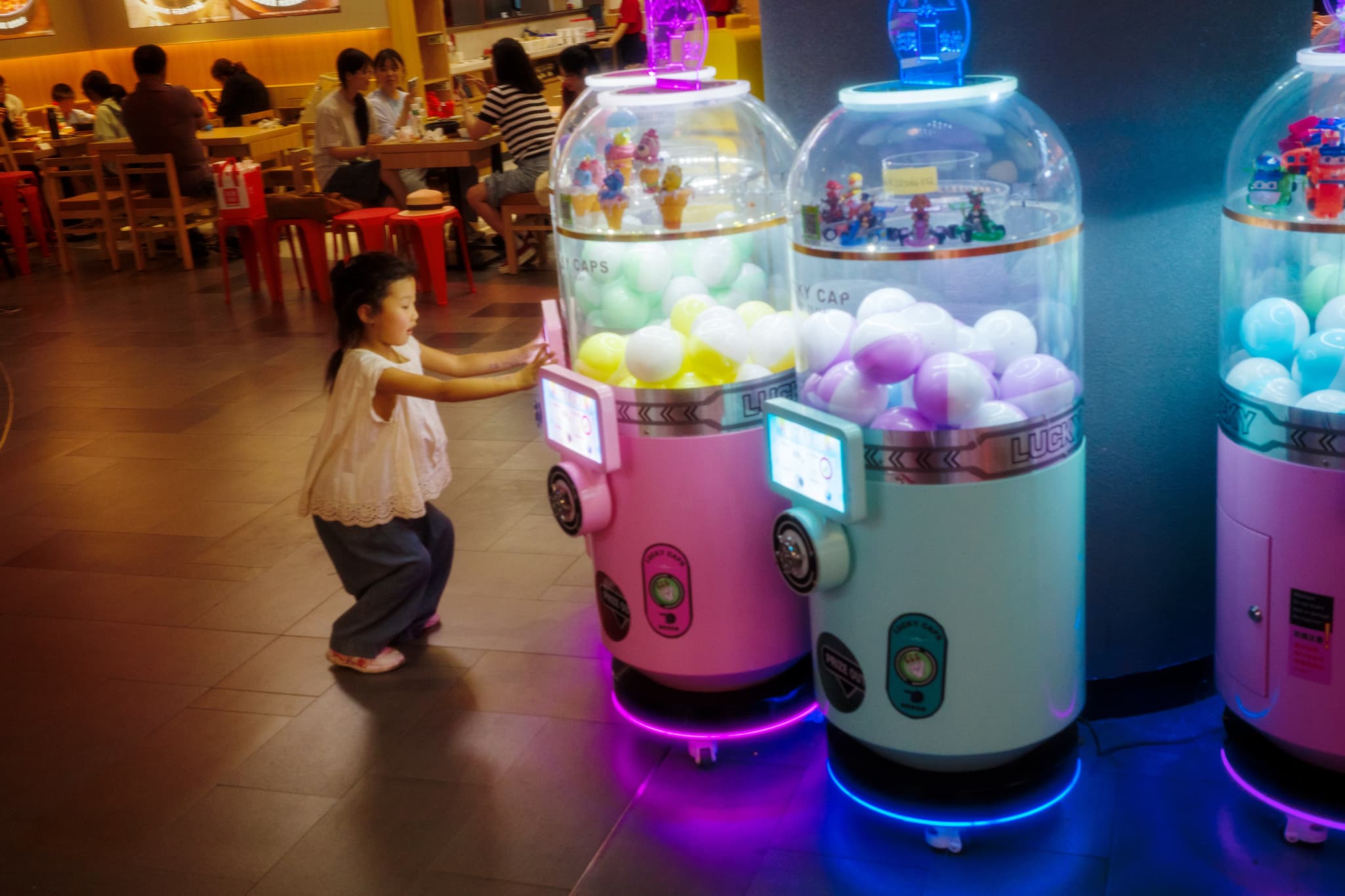 A young girl interacts with colorful claw machines filled with toy balls in a brightly lit indoor setting