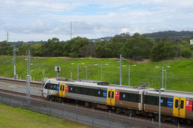 A commuter train travels along a multi-track railway through a green suburban landscape under a partly cloudy sky