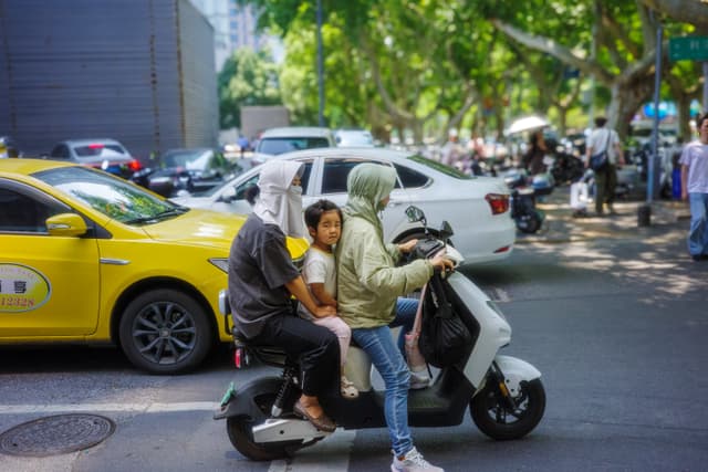 Three people riding a scooter on a busy urban street, surrounded by cars and trees