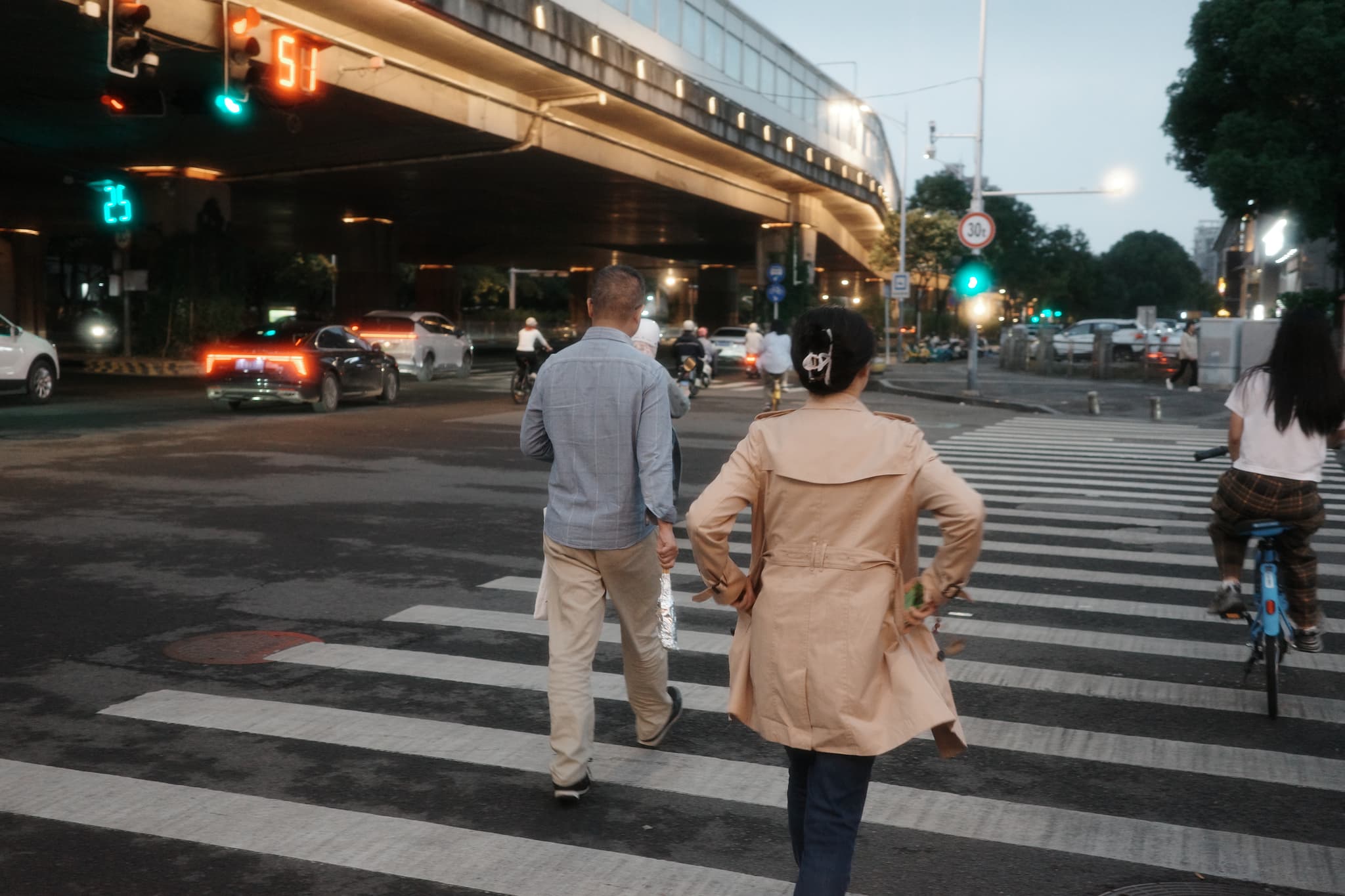 Pedestrians and a cyclist crossing a street at dusk near an overpass, with traffic lights and vehicles in the background