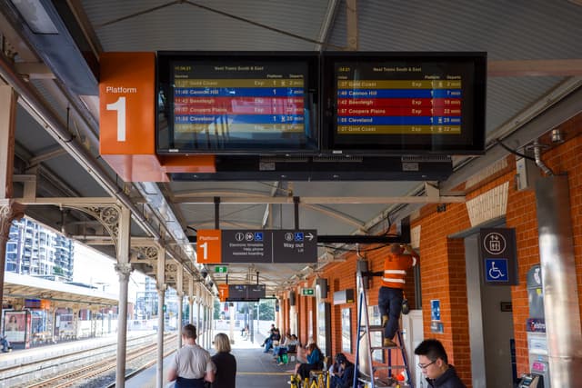 Train station platform with digital departure boards, passengers waiting, and orange signage under a covered roof