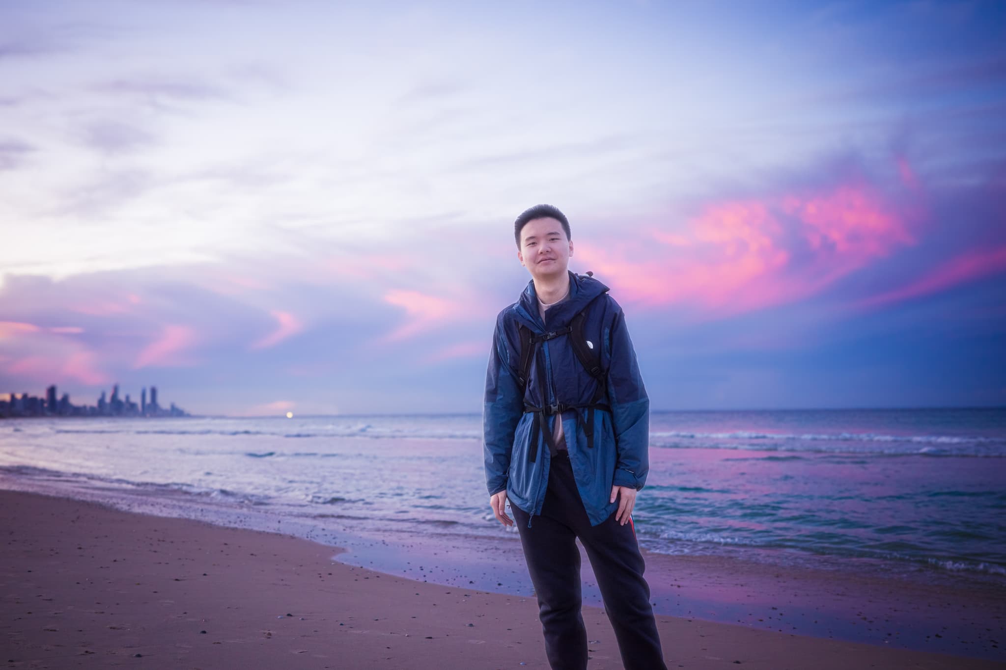 Person standing on a sandy beach near the waterline at dusk, wearing a backpack with colorful sunset clouds over the sea
