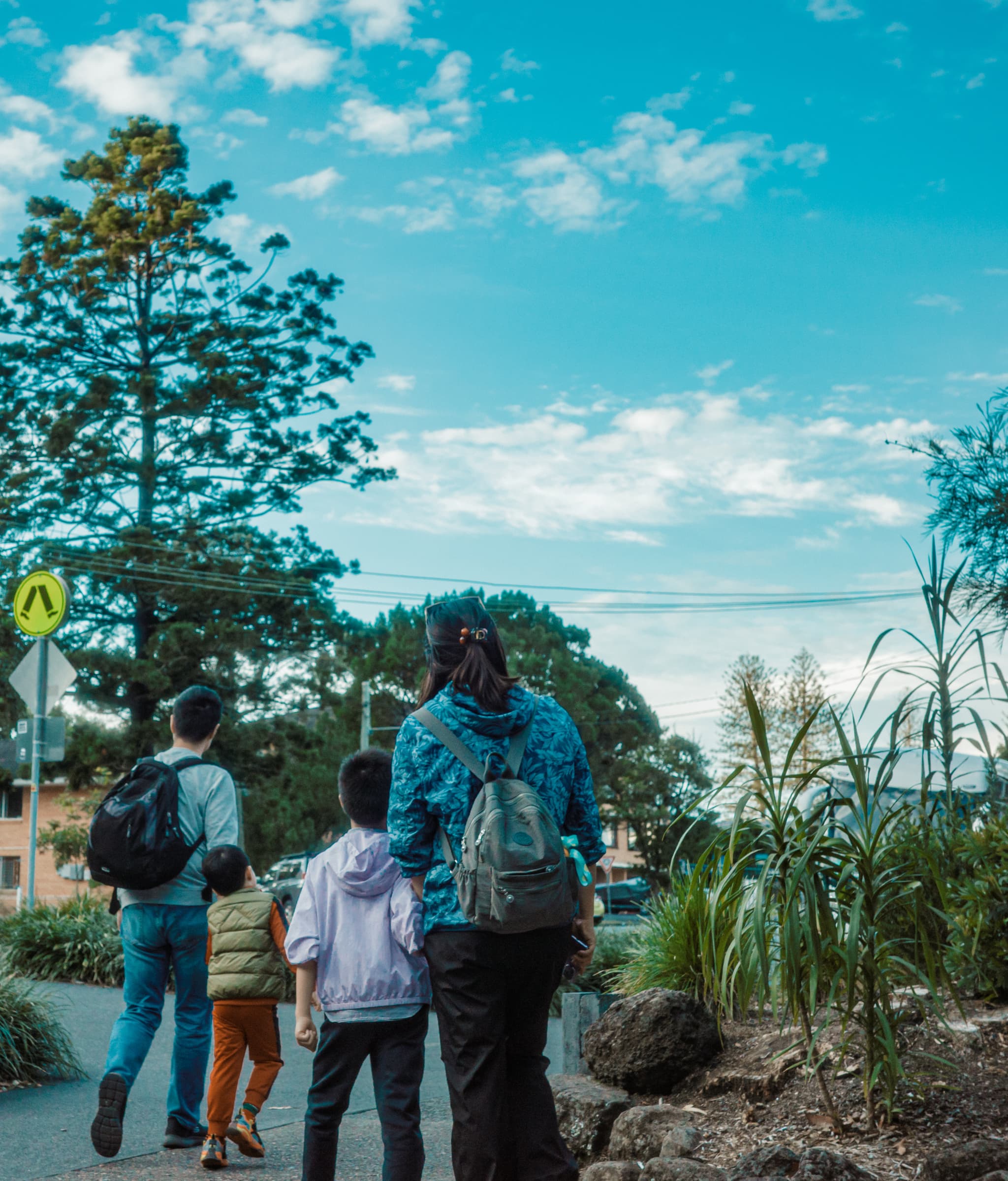 Three people walking along a roadside path near tall grasses and trees under a partly cloudy sky