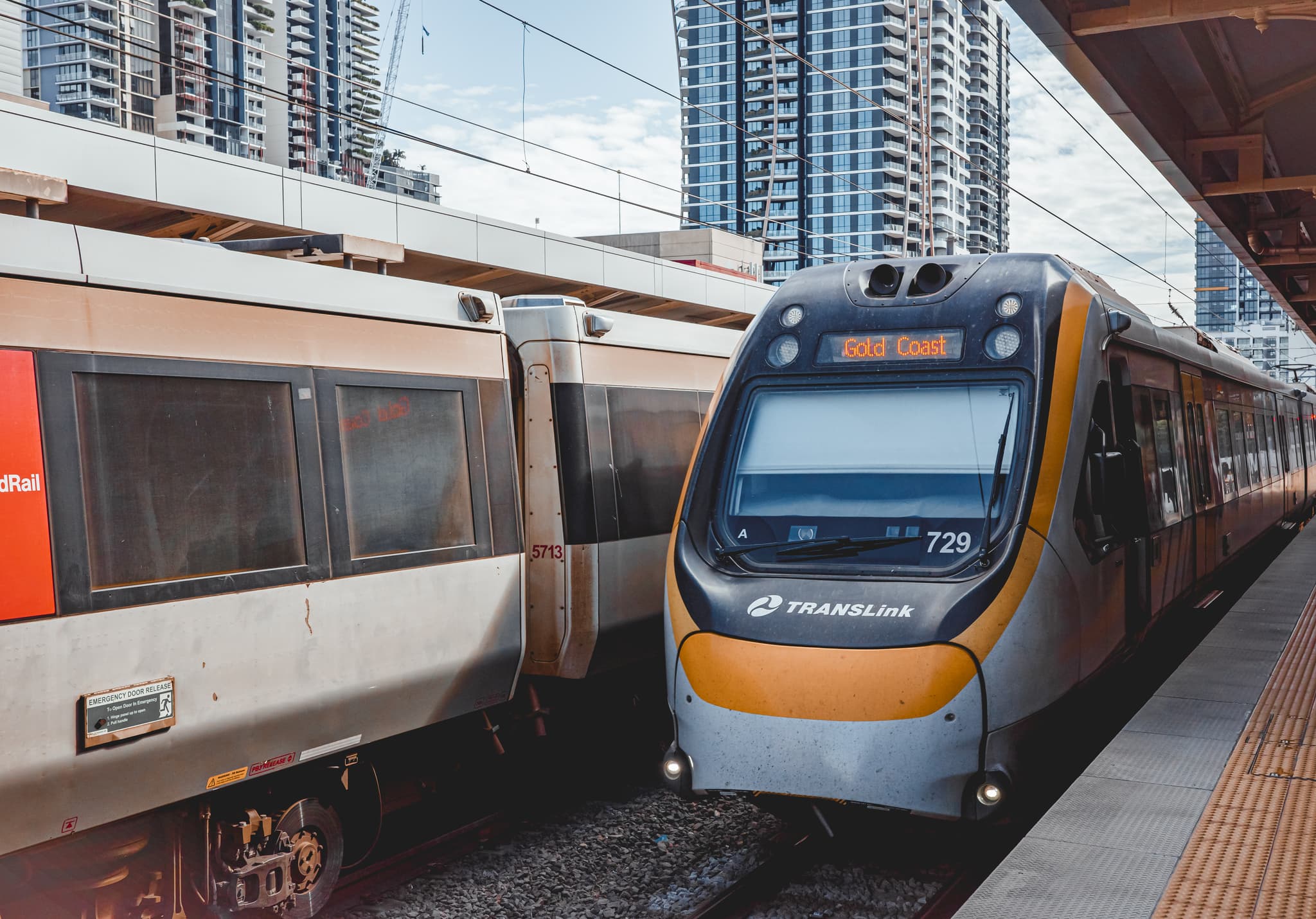 Two trains stopped side-by-side at a station platform, including a modern commuter train with a yellow-and-gray front under overhead lines