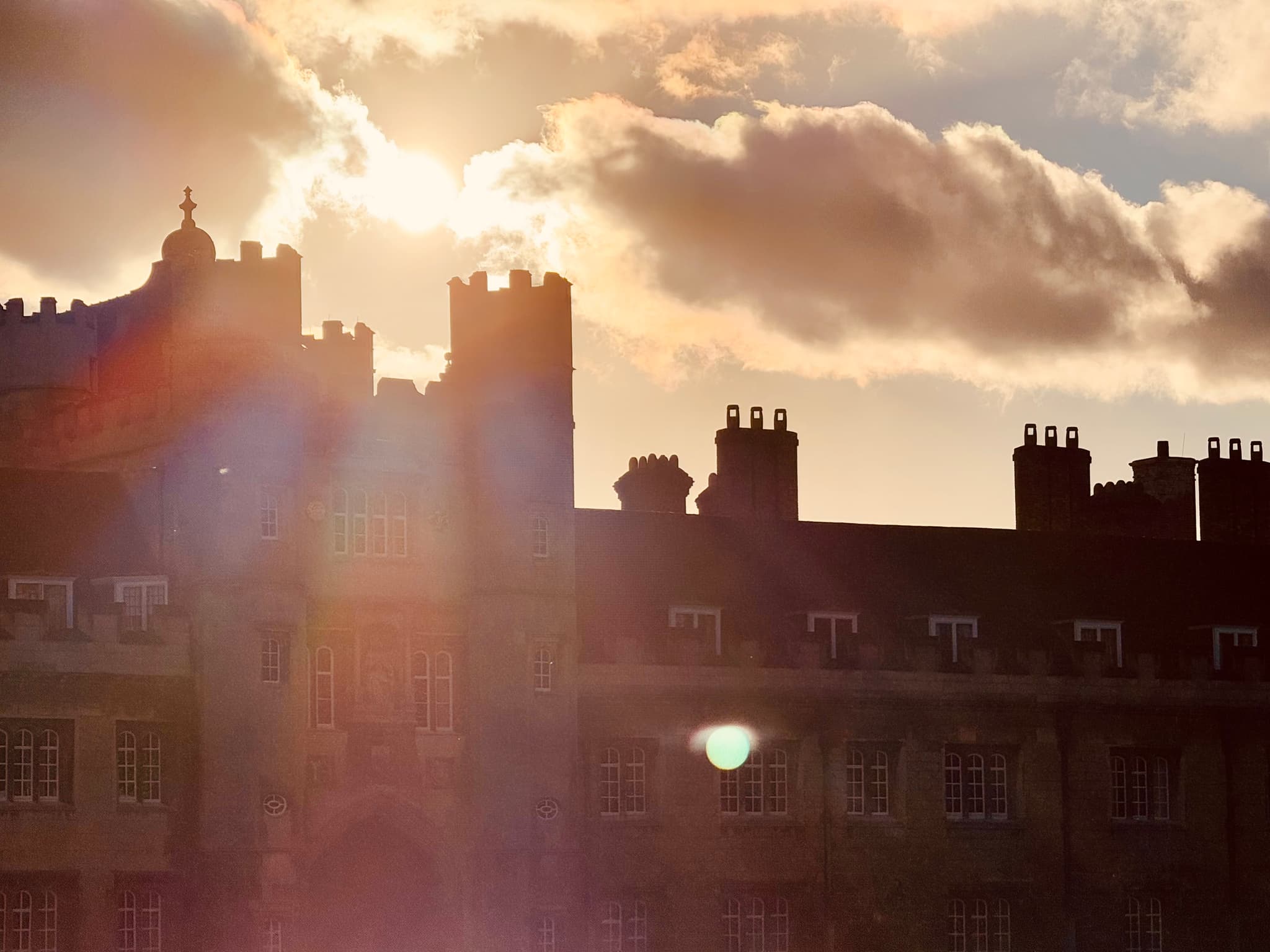 Sunset light flares behind silhouetted rooftops and chimneys beneath layered clouds