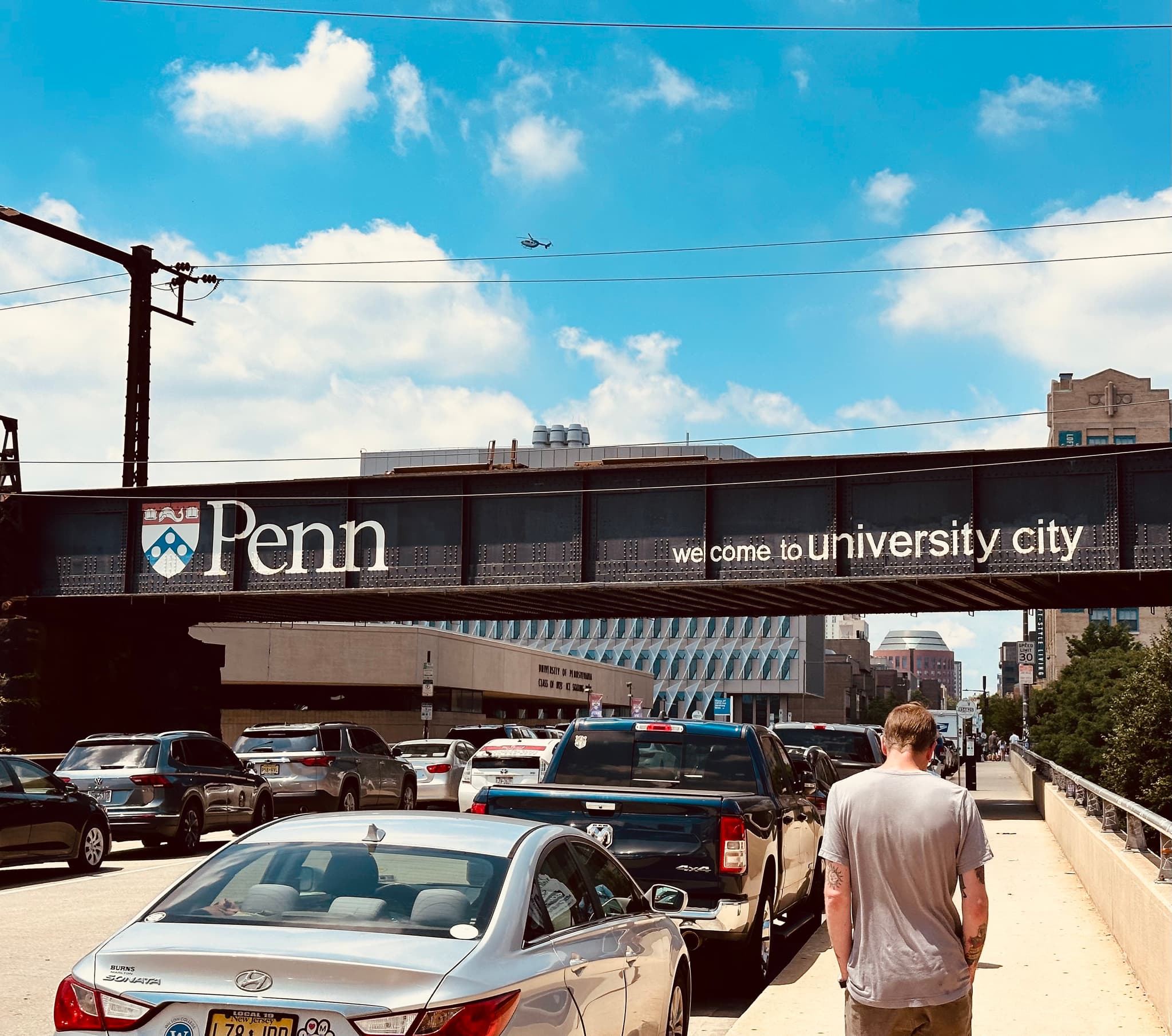 Pedestrian skybridge with a large Penn sign spanning a busy roadway with cars, a person walking, and university buildings in the background under a blue sky