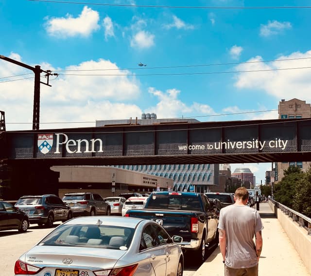 Pedestrian skybridge with a large Penn sign spanning a busy roadway with cars, a person walking, and university buildings in the background under a blue sky