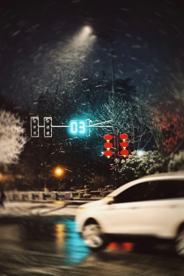 A nighttime scene with a traffic light displaying a red stop signal and a countdown timer set at 03 seconds Snowflakes are visible in the air, illuminated by a streetlight, and a blurred white car is in motion in the foreground