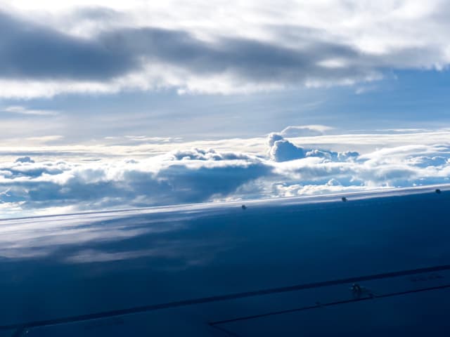 A serene skyscape with fluffy clouds and a clear blue sky, possibly taken from an airplane given the angle and the presence of what appears to be part of an aircraft's structure in the lower corner