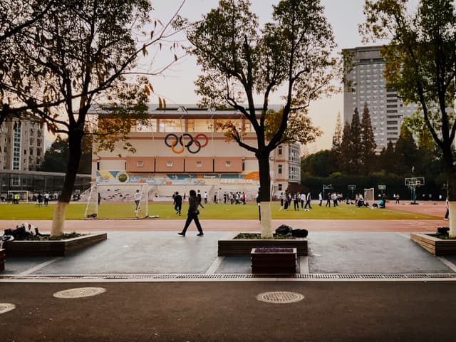 A sports field surrounded by trees, with a building displaying Olympic rings in the background and a person walking in the foreground