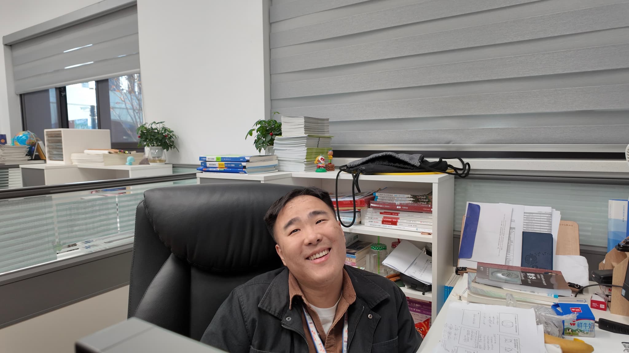 An office workspace with a desk, chair, shelves, documents, and various items, surrounded by windows and blinds