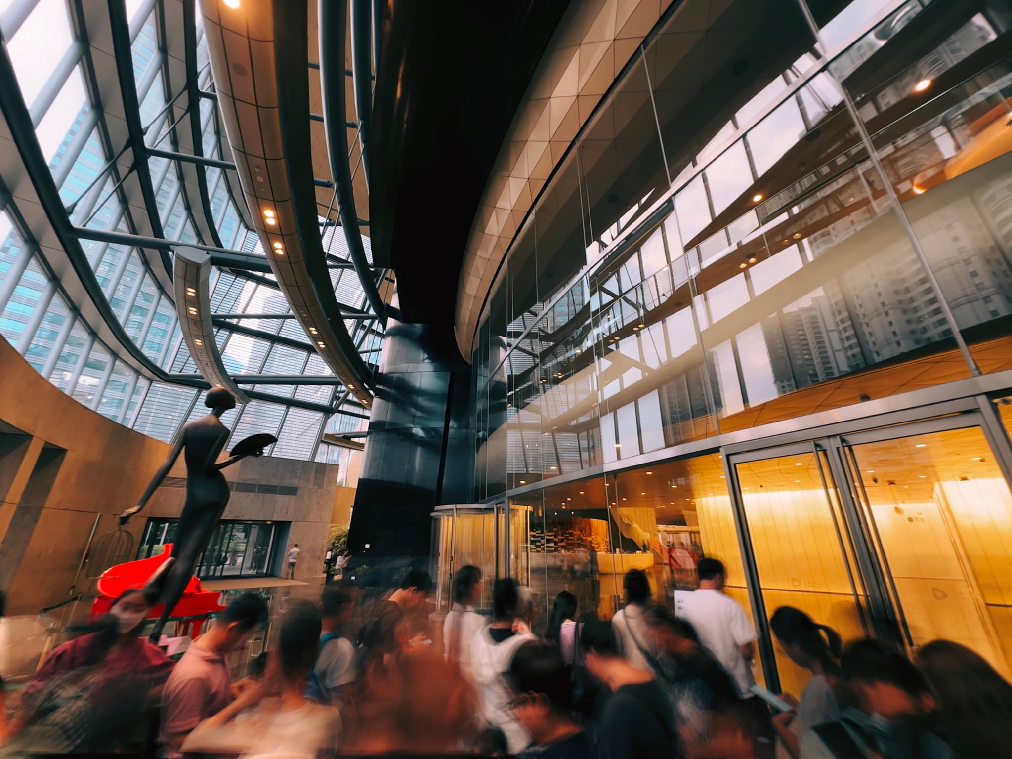 A bustling indoor scene with a crowd of people, possibly in a museum or gallery, with a curved architectural feature overhead and warm lighting from the surrounding windows and doors