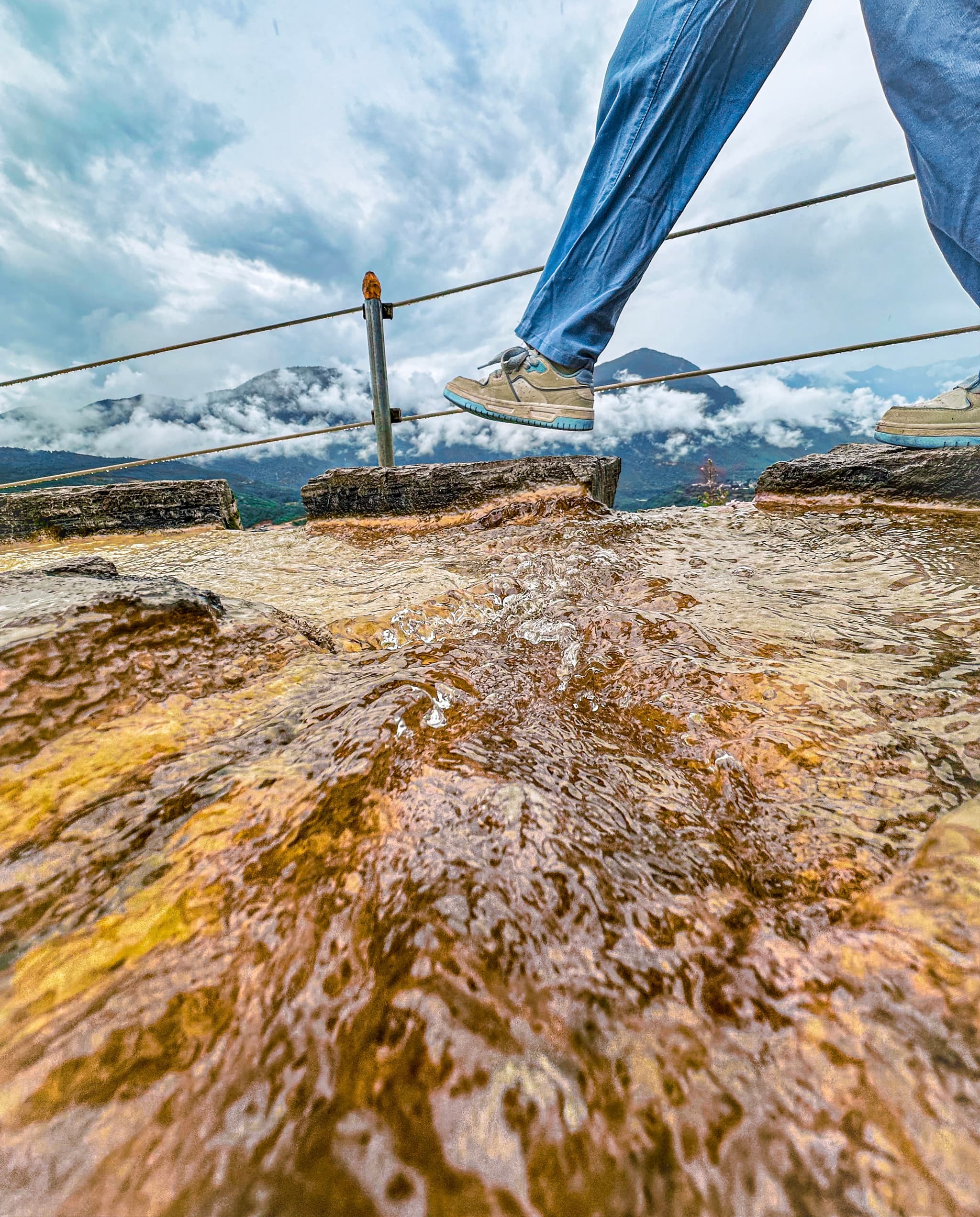 A person is walking across a wet, rocky surface with a rope handrail, against a backdrop of mountains and cloudy skies