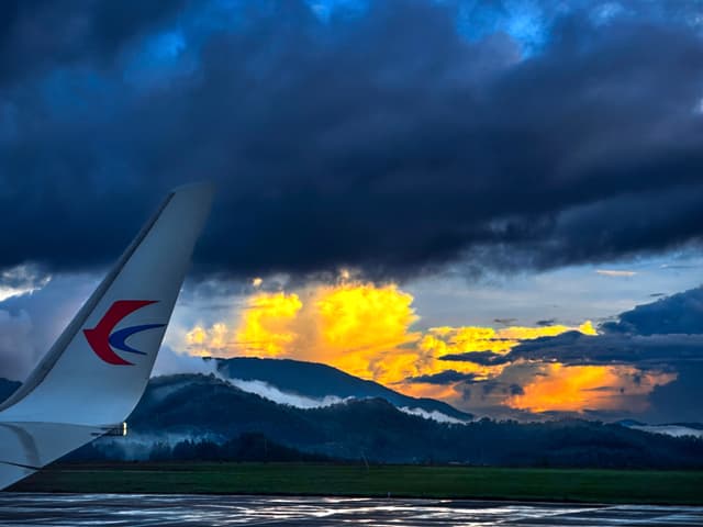 An airplane wing with a distinctive logo against a backdrop of a dramatic sunset with dark clouds and a mountainous horizon