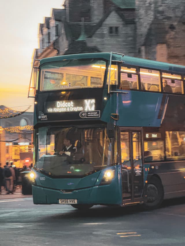 A double-decker bus labeled Didcot X2 driving through a town at dusk, with historic buildings in the background