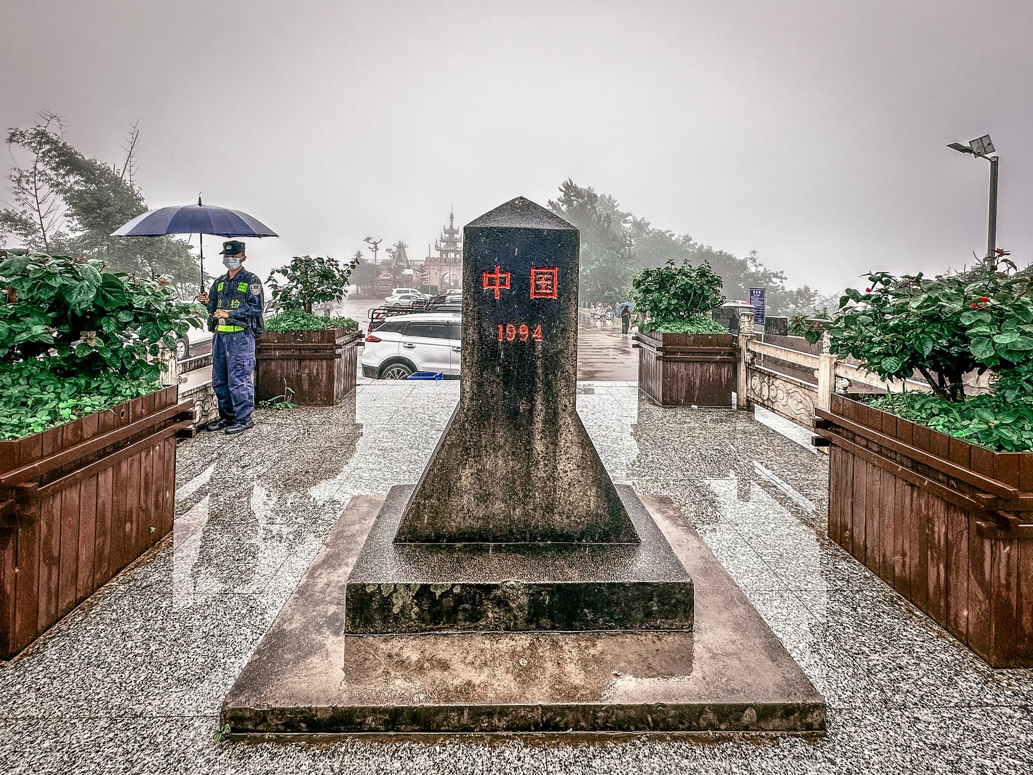 A rainy scene with a person holding an umbrella standing next to a monument with inscriptions, surrounded by greenery