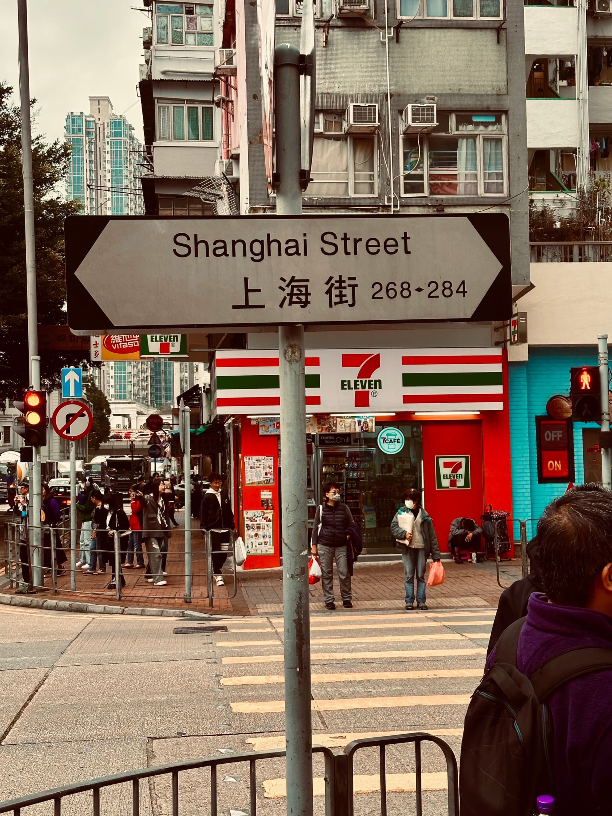 A street sign reading Shanghai Street with Chinese characters below, in an urban setting with pedestrians, a convenience store, and traffic lights in the background