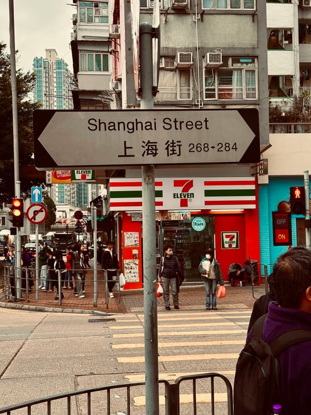 A street sign reading Shanghai Street with Chinese characters below, in an urban setting with pedestrians, a convenience store, and traffic lights in the background