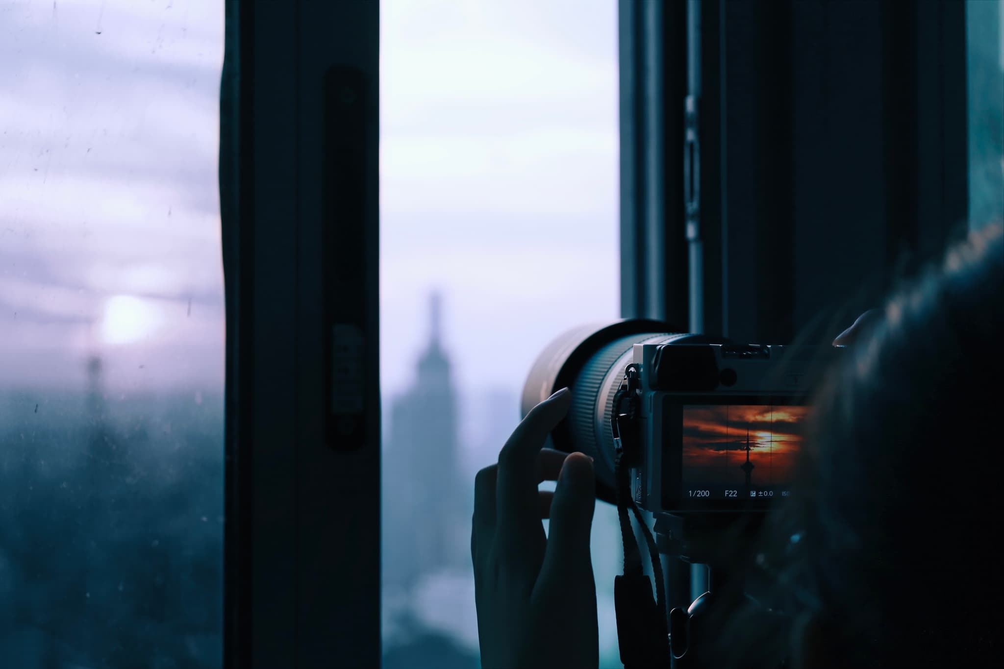 A person photographing a cityscape through a window during sunset, with a blurred building in the background