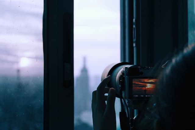A person photographing a cityscape through a window during sunset, with a blurred building in the background