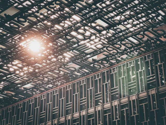 A complex metal framework forms the ceiling of a large space, with a single bright light source shining through