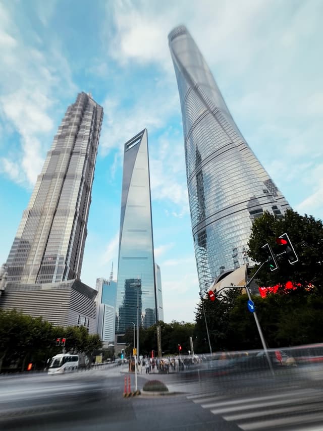 Skyscrapers reach towards a blue sky with scattered clouds, with the tallest one shrouded in a light mist at the top A city street with traffic lights and a pedestrian crossing is visible in the foreground