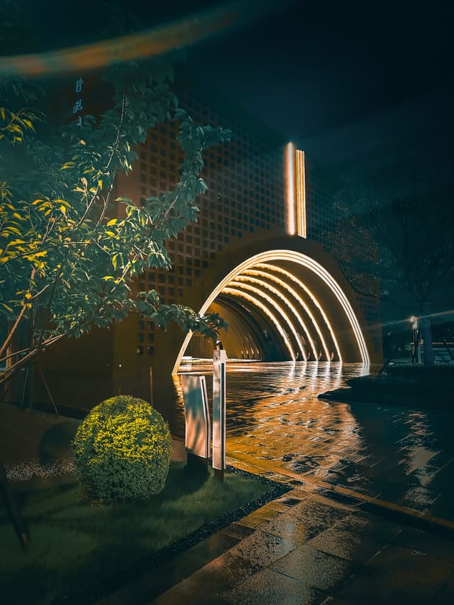 A nighttime scene with illuminated arches creating a tunnel effect, surrounded by lush greenery and a reflective surface, possibly water, on the ground The lighting casts a warm glow and highlights the architectural details of the surrounding buildings