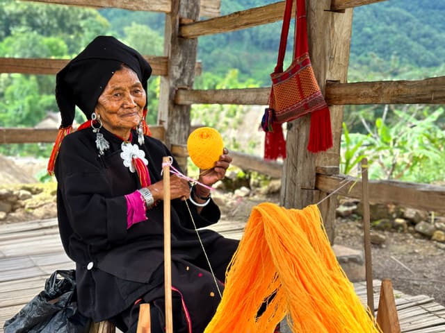 An elderly woman in traditional attire is smiling while winding bright orange yarn on a spindle, seated under a wooden shelter with lush greenery in the background