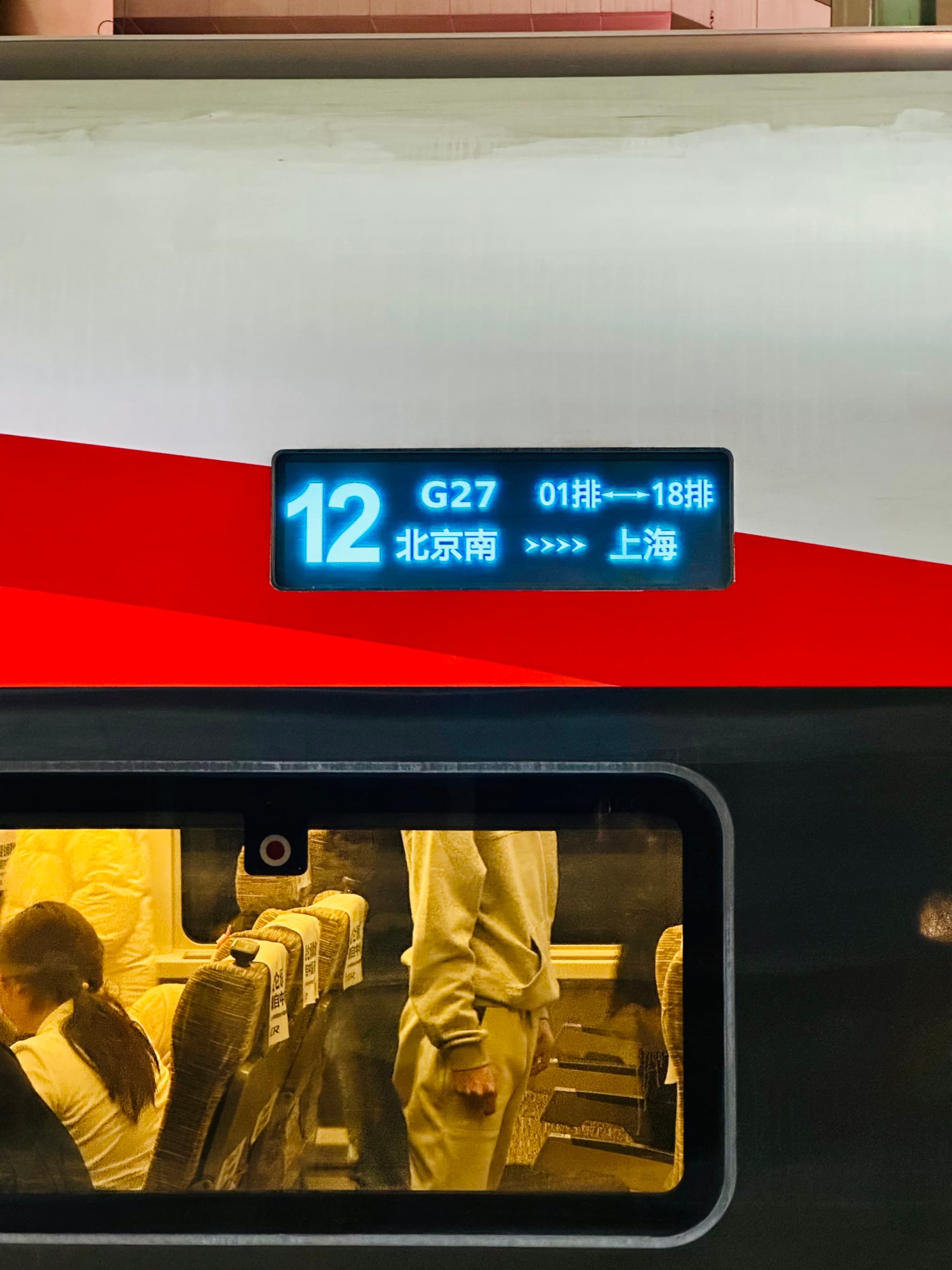 A train car with a digital display showing the number 12 and additional information, likely a train identifier or route information, above a window where passengers can be seen inside