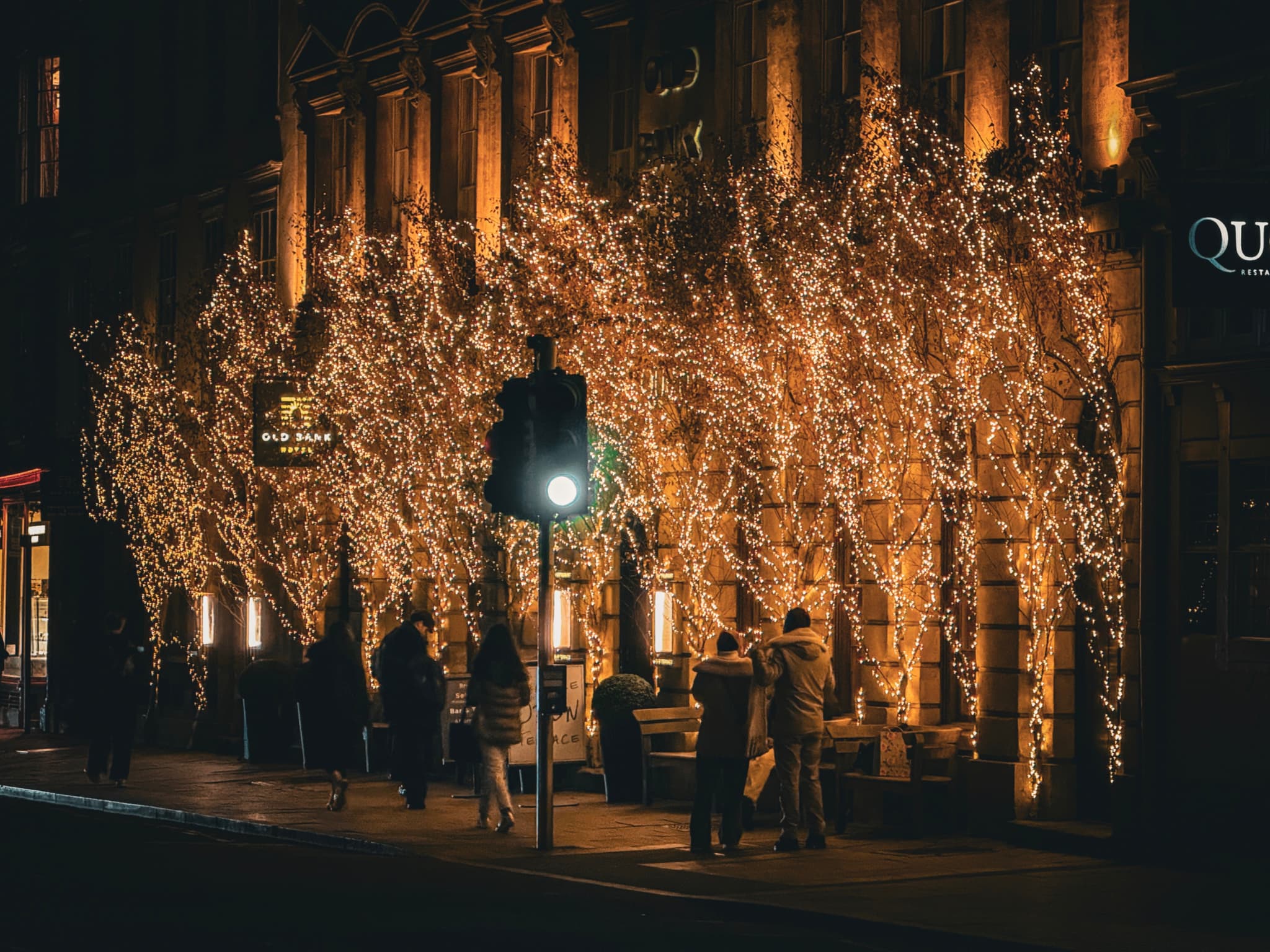 A warmly lit building adorned with glowing string lights, with people gathered outside and a traffic light in the foreground