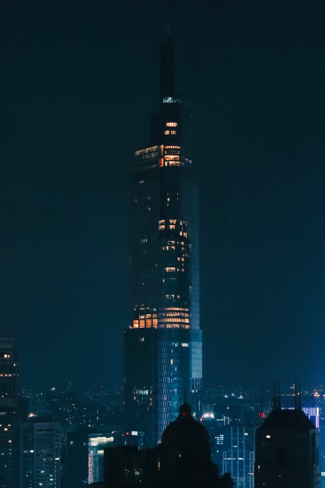 A tall, illuminated skyscraper stands against a dark night sky, surrounded by smaller buildings