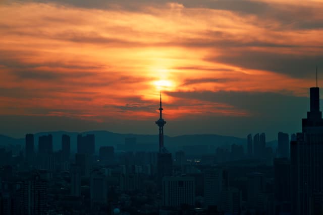 A city skyline at sunset with a prominent tower silhouetted against a vibrant orange and yellow sky