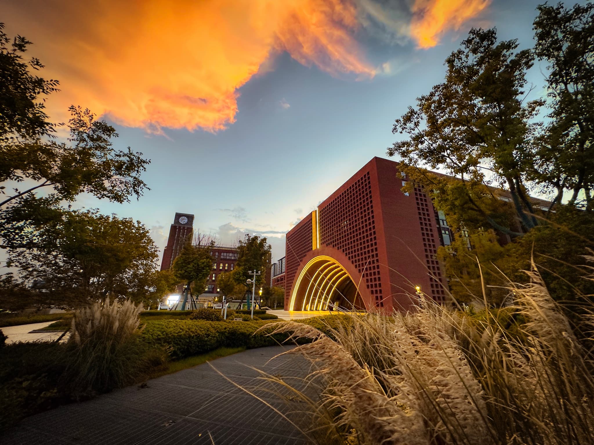 A modern building with a unique arched entrance and a red brick facade, set against a backdrop of a dramatic sky with orange clouds at dusk, surrounded by lush greenery and a winding path