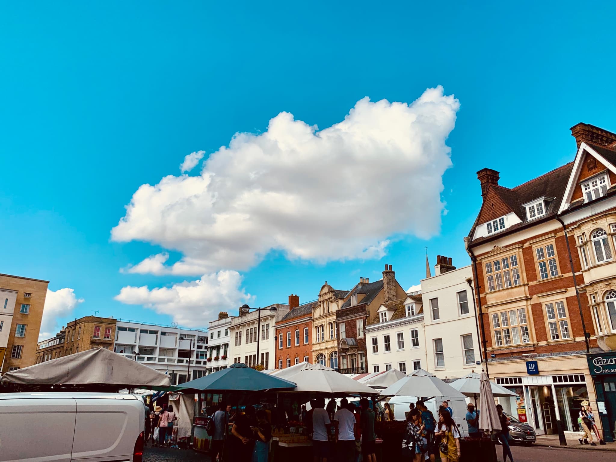 A bustling market square with tents and a crowd of people, framed by historic buildings under a blue sky with a prominent white cloud