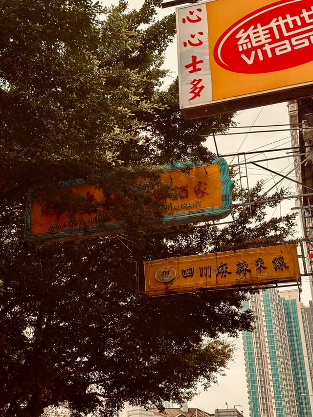 Vertical banners with Asian characters hang above a street, partially obscured by a tree, with a glimpse of a high-rise building in the background