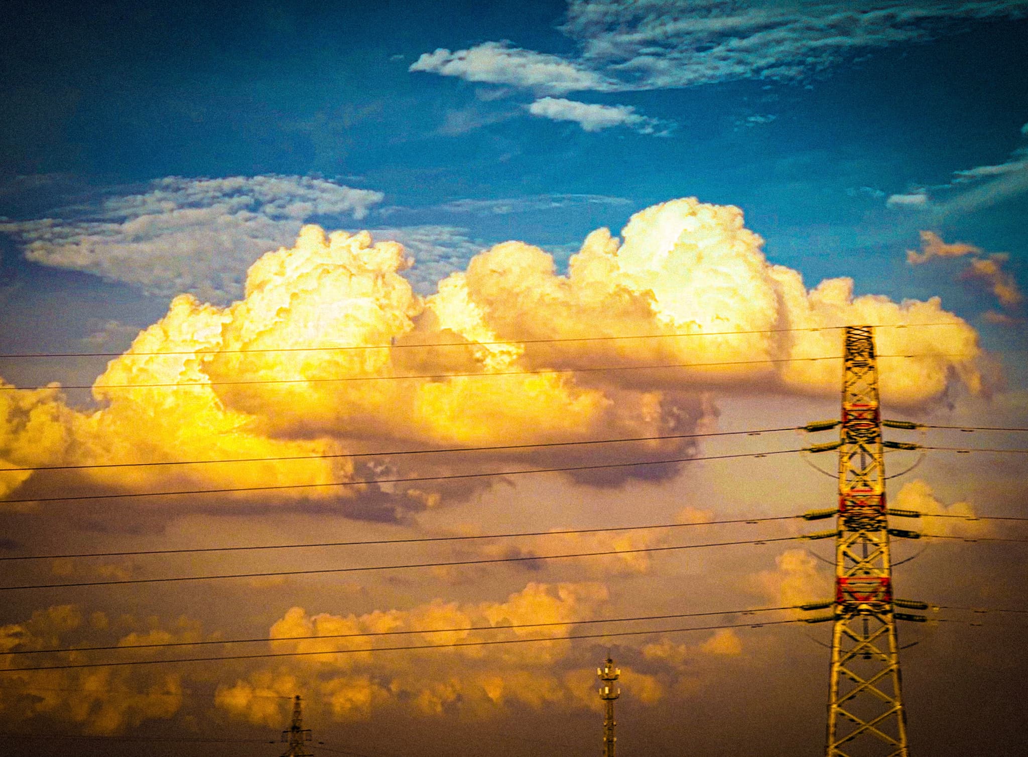 Cumulus clouds illuminated by golden sunlight with an electricity pylon in the foreground against a blue sky