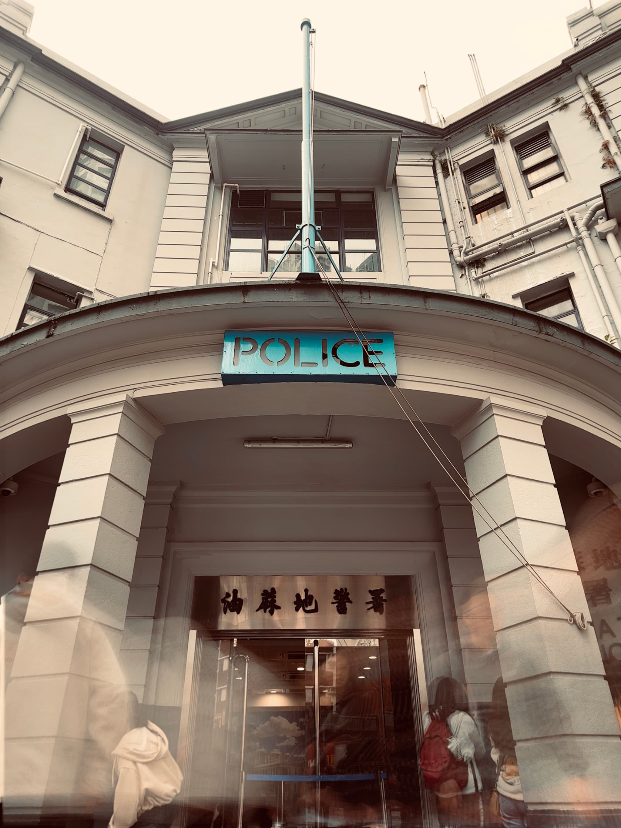 A police station entrance with a blue POLICE sign above the door, flanked by two flags on poles, and Asian characters on the door's glass