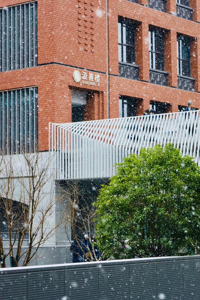 A red-brick building with modern architectural elements, surrounded by greenery, under falling snow
