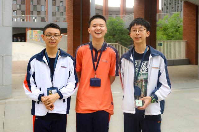 Three young men are standing together smiling, with one in the center slightly taller than the others They are dressed in casual and sporty attire, with the central figure wearing an orange shirt and a lanyard around his neck, and the others in jackets They appear to be in an outdoor setting with a building structure in the background.