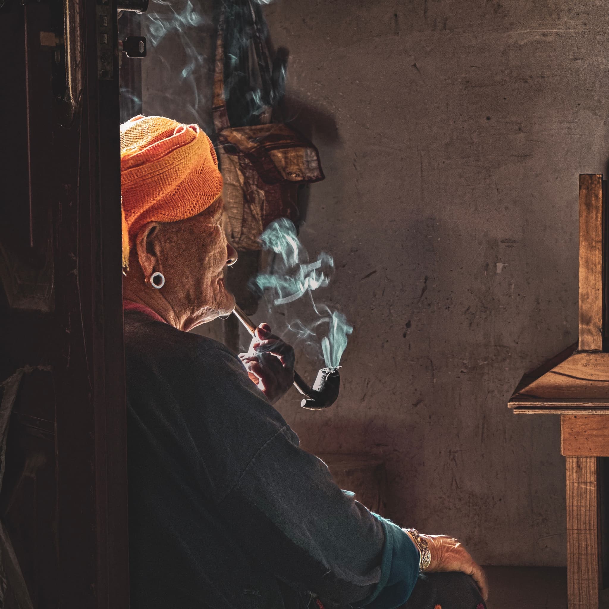 An elderly person wearing a headscarf is sitting and smoking a pipe, with smoke curling up in the air, in a room with subdued lighting