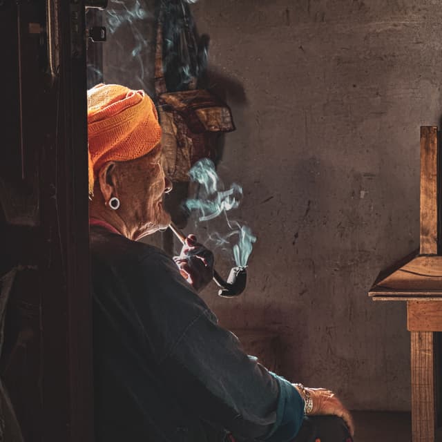 An elderly person wearing a headscarf is sitting and smoking a pipe, with smoke curling up in the air, in a room with subdued lighting