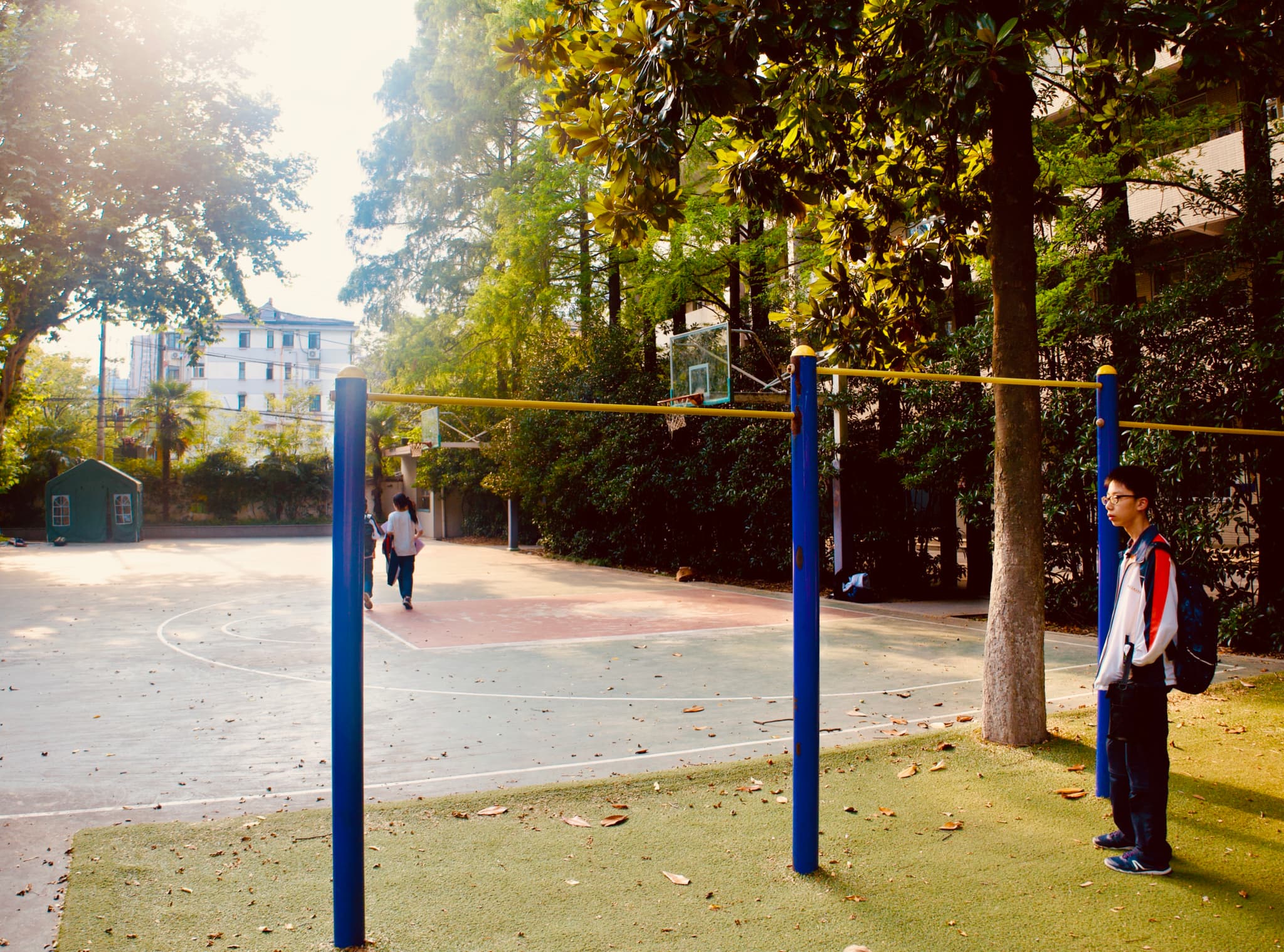 A schoolyard with a pull-up bar, surrounded by trees, where a student stands nearby and others walk in the background